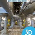 Das Bild zeigt den Innenraum eines Wasserwerks oder einer ähnlichen technischen Anlage. Es ist eine Perspektive von unten nach oben in einen Raum mit vielen großen Rohren und Behältern, die in einem regelmäßigen Muster angeordnet sind. Die Rohre sind silbergrau und verbinden die verschiedenen Behälter. Der Boden besteht aus großen, hellen Fliesen.  Im Vordergrund befindet sich ein Logo, das auf ein 50-jähriges Jubiläum des „Rhein Kreis Neuss“ hinweist.  Das Bild wirkt technisch und sachlich. © Kreiswerke Grevenbroich GmbH