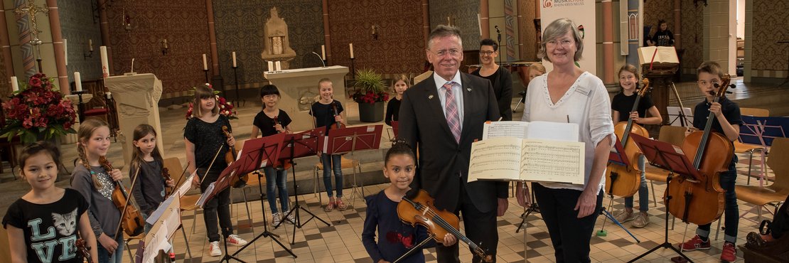 Mehrere Kinder stehen im Halbkreis mit Musikinstrumenten in einer Kirche. Im Vordergrund stehen zwei Erwachsene.