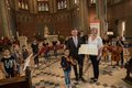 Mehrere Kinder stehen im Halbkreis mit Musikinstrumenten in einer Kirche. Im Vordergrund stehen zwei Erwachsene.