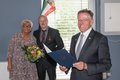 Landrat Hans-Jürgen Petrauschke hält die Verleihungsurkunde in den Händen. Versetzt dahinter stehen Erik Heinen, mit Bundesverdienstkreuz am Revers, und Ehefrau Michaela mit einem Strauß Blumen in der Hand. 