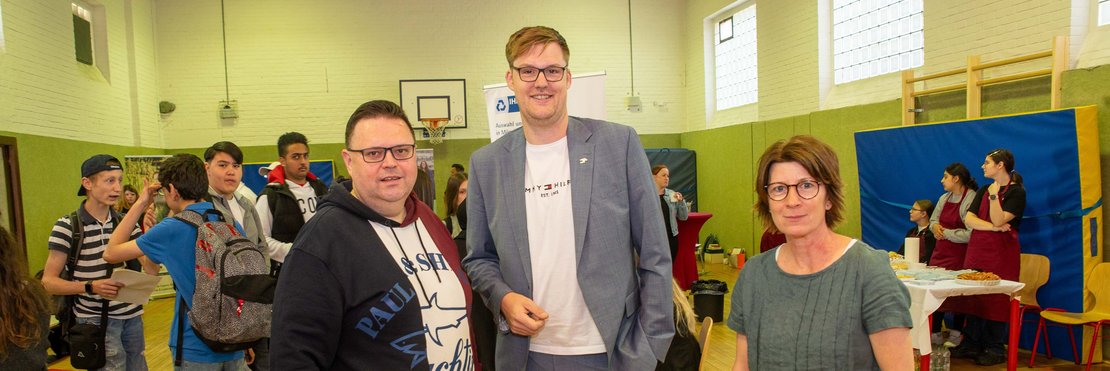 Eine Frau und zwei Männer stehen in einer Turnhalle und schauen in die Kamera. Im Hintergrung stehen Schüler an verschiedenen Ständen.
