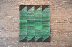 Wandbehang aus vier senkrechten Streifen, die wiederum aus kleinen waagerechten grünen Streifen bestehen vor einer Backsteinwand.