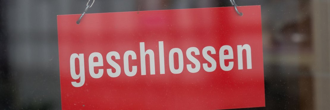 An einer Glastür hängendes rotes Schild mit weißem Schriftzug "geschlossen"