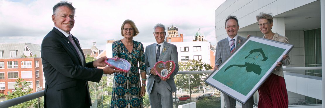 Landrat Hans-Jürgen Petrauschke, das Neusser Schützenkönigspaar Kurt und Beate Koenemann, Schützenpräsident Martin Flecken und Ehefrau Gabi stehen mit Abständen auf dem Balkon im Kreishaus Neuss. Herr und Frau Flecken halten einen Bilderrahmen in den Händen auf dem ein künstlerisch gestalteter Quirinuskopf mit einer goldenen Träne zu sehen ist. Landrat Petrauschke übergibt der Schützenkönigin Lebkuchenherzen. Im Hintergrund ist die Neusser Innenstadt und das Quirinusmünster zu sehen. 