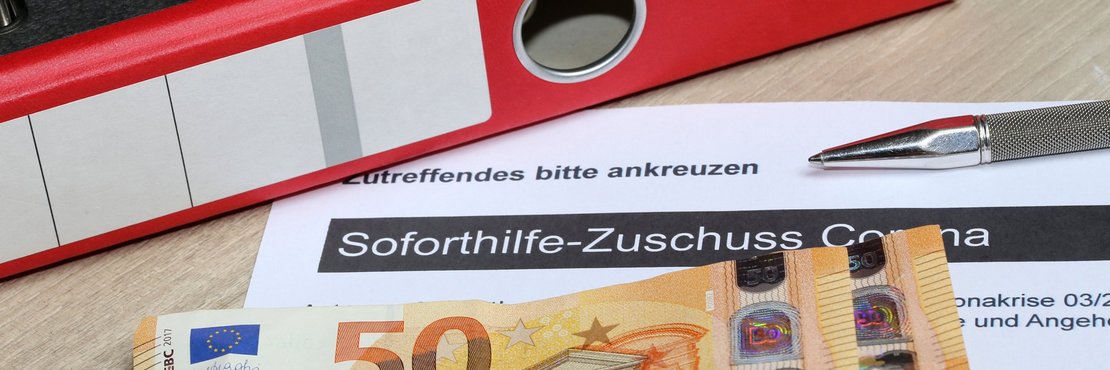 Geldscheine liegen auf Corona-Soforthilfe-Antrag vor einem roten Aktenordner