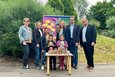 Das Bild zeigt eine Gruppe von Erwachsenen und Kindern vor einem Schaukasten mit Informationen über Wild- und Honigbienen. Die Kinder halten jeweils eine Dose mit Saatgut oder ähnlichem in der Hand. Die Erwachsenen stehen hinter den Kindern und scheinen mit dem Projekt etwas zu tun zu haben. Es ist wahrscheinlich eine Aktion zum Schutz der Bienen.  Der Hintergrund besteht aus Grünflächen und Bäumen. Die Atmosphäre wirkt freundlich und positiv.