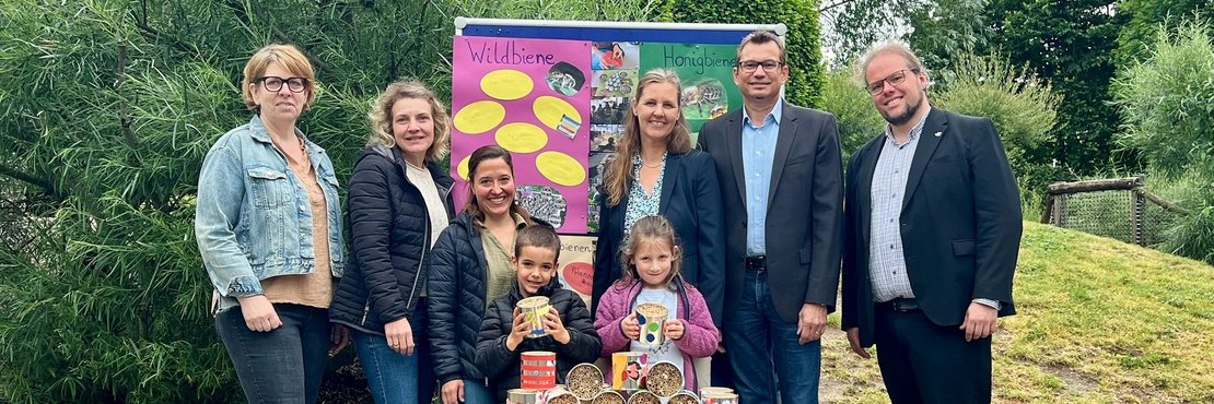 Das Bild zeigt eine Gruppe von Erwachsenen und Kindern vor einem Schaukasten mit Informationen über Wild- und Honigbienen. Die Kinder halten jeweils eine Dose mit Saatgut oder ähnlichem in der Hand. Die Erwachsenen stehen hinter den Kindern und scheinen mit dem Projekt etwas zu tun zu haben. Es ist wahrscheinlich eine Aktion zum Schutz der Bienen.  Der Hintergrund besteht aus Grünflächen und Bäumen. Die Atmosphäre wirkt freundlich und positiv.
