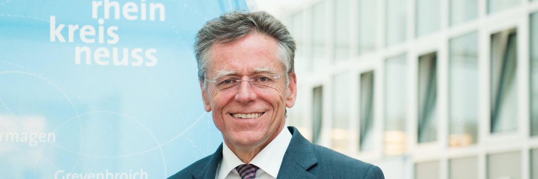 Landrat Hans-Jürgen Petrauschke steht vor einer blauen Kreisfahne und lächelt