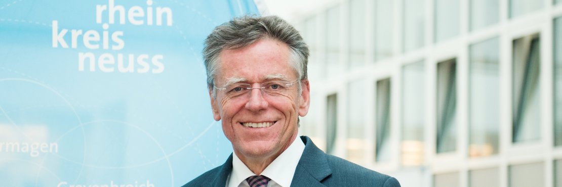 Landrat Hans-Jürgen Petrauschke steht vor einer blauen Kreisfahne und lächelt