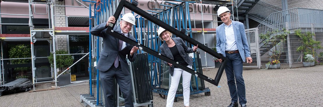 Landrat Hans-Jürgen Petrauschke, Christina Grebe und Johannes Gillrath mit Schutzhelmen halten ein Fenster vor einem Schulgebäude mit Baugerüst.