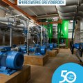 Das Bild zeigt eine Maschinenhalle mit mehreren blauen Pumpen und einem komplexen System aus grauen Rohren und Ventilen. Die Pumpen stehen auf quadratischen Sockeln aus braunen Fliesen. Im Hintergrund befinden sich zwei große grüne Behälter. Die gesamte Szenerie wirkt industriell und technisch.  Das Bild fokussiert auf die technische Anlage und wirkt sauber und ordentlich. © Kreiswerke Grevenbroich GmbH