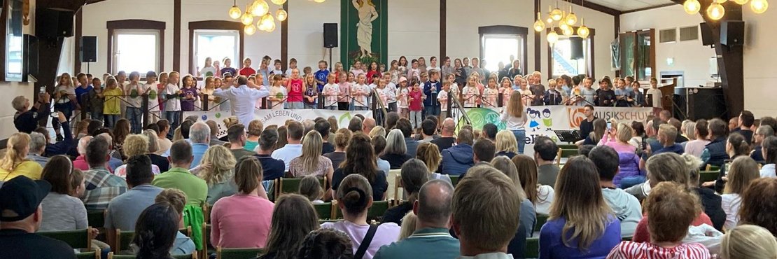 Konzertsaal mit vielen Gästen und einer großen Gruppe Kinder, die auf einer Bühne stehen und singen. Davor ein Lehrer, der dirigiert.