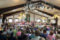 Konzertsaal mit vielen Gästen und einer großen Gruppe Kinder, die auf einer Bühne stehen und singen. Davor ein Lehrer, der dirigiert.